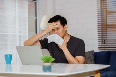 Handsome Asian man sitting at home with sickness, fever, stuffy nose, sneezing, runny nose, holding toilet paper, wiping fluid on his nose.の写真素材