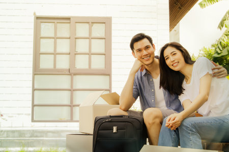 Young couple rejoiced, feeling excited and happy together while sitting in wooden chairs ready to move their bags, clothes and boxes into their new home behind.の写真素材