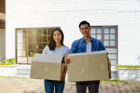 Moving into a new home : Young couple standing outside of the house, holding a cardboard box, are happy to move the cardboard boxes.の写真素材