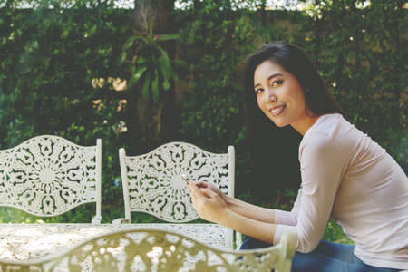 Asian woman sitting on a white alloy table relaxing in the morning in the shady garden, her daily life is on her cell phone to sit comfortably in her spare time.の写真素材