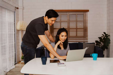 Asian couples work online together to discuss online trade documents and do their work at home on their laptops.Look at the laptop: couples plan their business and work from home.の写真素材