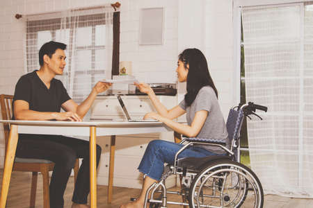 Disabled beautiful woman sitting on a wheelchair working with the male boyfriend in the home office, helping to deliver papers to the girlfriend who must help each other on the desk with understandingの写真素材