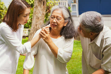 Old woman walking outside, feeling exhausted, fainted, and supported by her husband and daughter, used a balm to help her breathe better, showing good care.の写真素材
