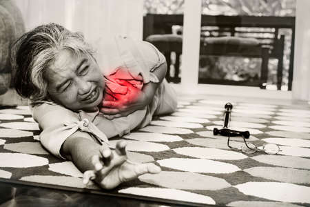 Black and white photo elderly Asian senior woman alone in the home, suffering from a heart attack, falls to the ground, glasses and a cane slip off in pain, seeking help.の写真素材