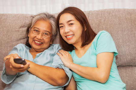 Portrait asian family happy sitting together on sofa at home : An elderly mother turns years and a daughter in the living room, Use the remote to sit and watch TV during happy moments.の写真素材