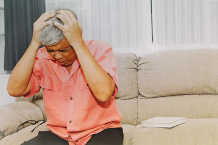 Senior man sat on the couch at home, getting dizzy and headaches during too long reading, causing stress.の写真素材