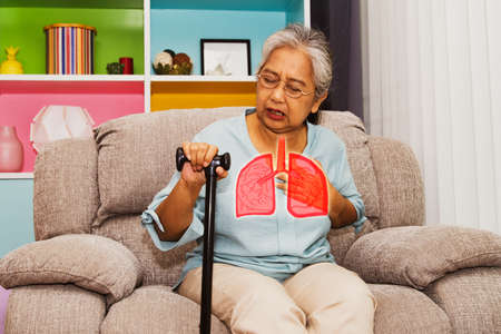 Elderly Asian woman suffers from respiratory pneumonia : Elderly woman sitting on a sofa is unwell, sick with influenza, has symptoms of depression, rapid breathing and chest pain.の写真素材
