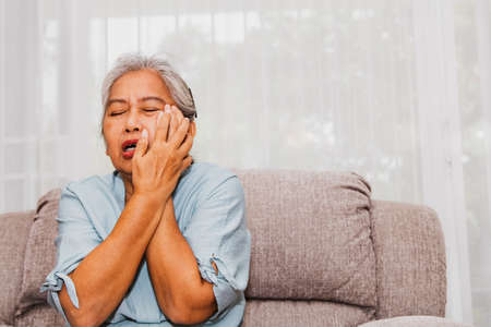 An elderly woman with toothache from inflammation of the gums, tooth decay, oral ulcers, pain until suffering, sits on the sofa with her hand holding her mouth, crying.の写真素材