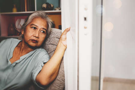 Elderly Asian woman sits at home to keep her health safe from the virus. She looked at the outside window, worried about the serious outbreak : Home to reduce the risk of infectionの写真素材