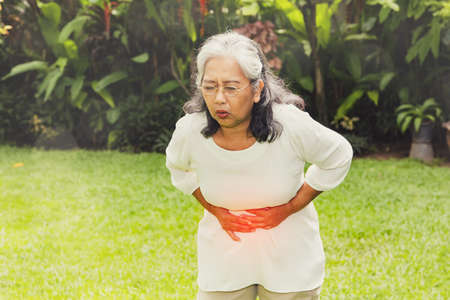 Elderly Asian woman has gastrointestinal problems, walking, abdominal pain, her stomach is bent, her hand touches her stomach, the pain makes her unable to walk : Retired women have health problems.の写真素材