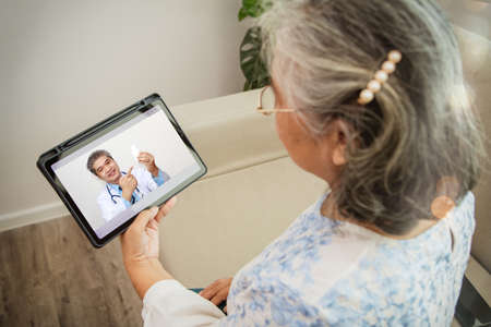 Top view elderly woman is making a video call with a medical professional while at home : Video conferencing with a consultant doctor on a digital tablet. Sick old woman in online consultation.の写真素材