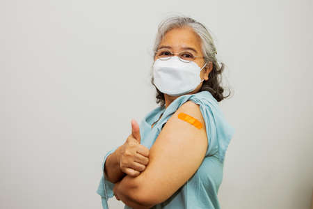 Pelderly asian elderly woman wearing mask received virus vaccination on arm and covered with germ-contaminating antimicrobial plaster is in good health showing excellent thumb upの写真素材