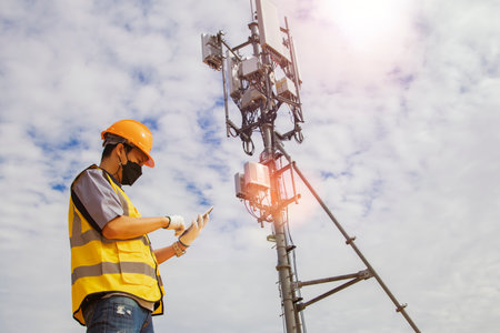 Asian male worker engineer wearing a helmet and safety goggles uses a smartphone to field work near a telecommunication tower controlling cellular electrical installations.の写真素材