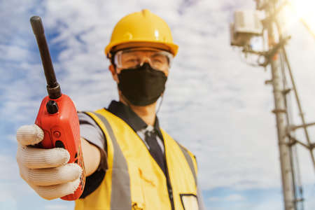 Male worker engineer wearing a helmet and safety goggles uses a walkie-talkie to do fieldwork on a telecommunication tower controlling the installation of cellular electrical equipment.の写真素材