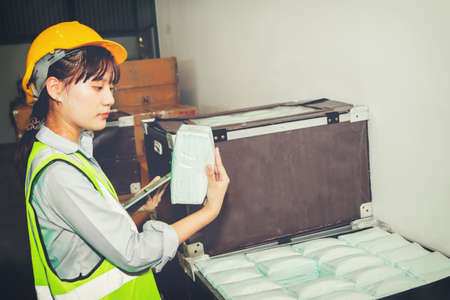 Female worker at an industrial factory producing face masks holding laptops and packed masks stands to inspect the orderliness of masks in boxes to be exported for sale.の写真素材