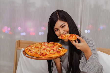 Beautiful Asian girl eats pizza gluttony is dinner, eaten before bedtime, eating in bed in her house with pleasure : Woman orders pizza online for home delivery during Covit-19 epidemicの写真素材