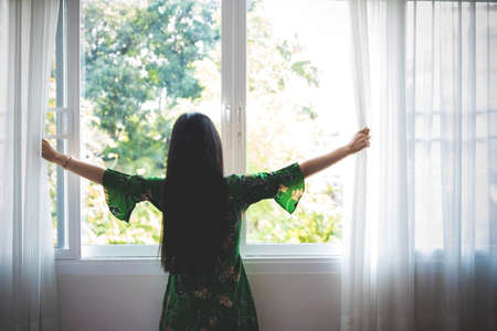 Sunny day : The woman stood, turned back, opened a white cloth in the room to receive a warm, bright morning light : Soft toneの写真素材