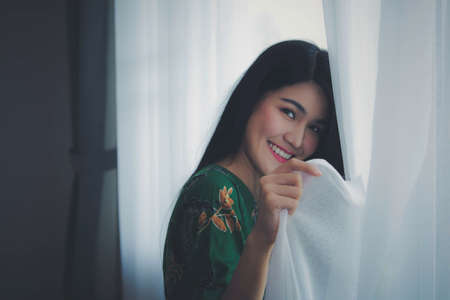 Portrait of an Asian woman, long hair, standing, opening a white curtain on the window, sending eyes and smiles with the morning joyの写真素材