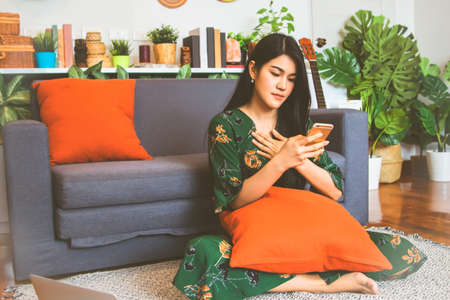 Asian woman with long hair wearing a green skirt, using a smartphone, looking at the phone, anxious gesture, sitting inside the living room.の写真素材