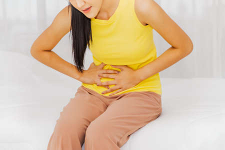 Women with abdominal pain and health care concept : Young beautiful woman touches her stomach, suffers lower abdominal pain, cystitis, she sits cramped in bed in the bedroomの写真素材