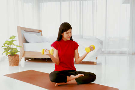 Home happiness concept : Young Asian woman with a laid back outfit, does a light workout with a small dumbbell in her clean bedroom in her home.の写真素材