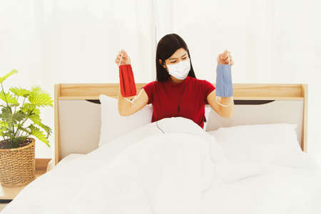 Concept new normal : Take care of the health of a beautiful Asian woman wearing a red shirt, wear a mask, sit on a bed, hold a new cotton mask to change the old mask that has been used for a long timeの写真素材