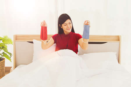 Normal new concepts:Portrait of a beautiful Asian woman wearing a red shirt, smiling comfortably on her bed at home, carrying a red and blue cloth mask suitable for traveling to work.の写真素材