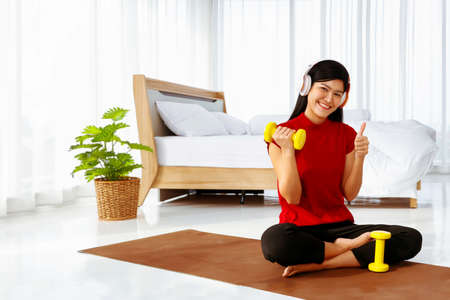 So happy, Young asian woman with a laid back outfit, does a light relay workout with music and lifts a small bright yellow dumbbell in her clean bedroom in a wonderful home.の写真素材