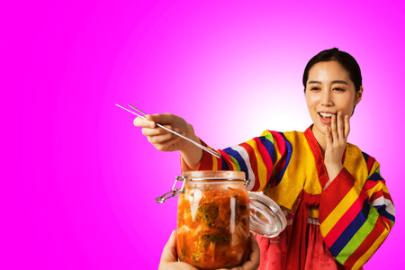 Beautiful young woman wear Korean traditional clothes, hanbok, taste kimchi in a glass jar and use chopsticks to eat in a good moodの写真素材