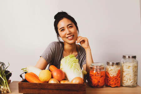 Happiness of an young asian woman preparing fermented food and eating kimchi in a glass jar with various kinds of fresh vegetables for making kimchi on a wooden table.の写真素材