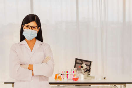 Female scientist wearing a mask stands across her arms in a chemical laboratory with science lab equipment, and she prepares for a test.の写真素材