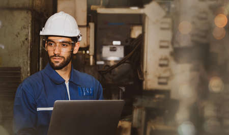 Professional handsome male engineer in blue uniform and helmet working on a laptop computer in the indoor field of a mechanical factory.の写真素材