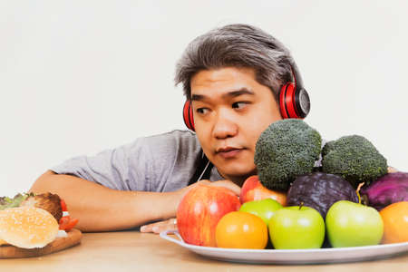 Fat Asian man trying to lose weight, hesitant to eat healthy fruits and vegetables : Man who glanced at junk food with contemplation decided not to eat the hamburger that was lying on the table.の写真素材