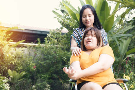 Female patients wearing yellow shirts are paralyzed, limb weakness, sitting on a wheelchair, are closely monitored by a caring female friend who takes a tour of the garden to relax : Selective Focusの写真素材