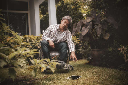 Handicapped Asian man in a wheelchair with health problems tries to save himself from a laptop that falls on the floor.の写真素材