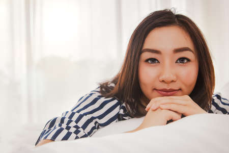 Asian teen girl lazing on the bed looking at the cute camera smiling brightly in the morning on a clean white bed at home.の写真素材