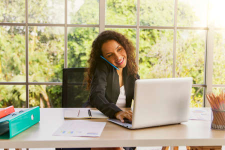 Portrait of a hospitable African American business woman talking on a mobile phone with a customer and typing a message on a keyboard. Save data to her laptop in the modern office with a smile.の写真素材
