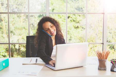 Portrait of an African American businesswoman talking on a mobile phone with a customer and typing messages on a keyboard, recording information on her laptop in a modern office with a smile.の写真素材
