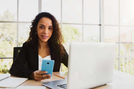 Portrait Business woman African American sitting comfortably holding a mobile phone using a laptop in a modern office.の写真素材