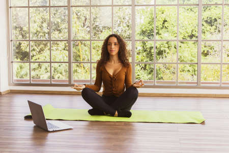 Healthy beautiful Young hispanic womanWearing sexy clothes showing breasts practice yoga practice teaching techniques on laptop calm mind meditation Sit on yoga mat in glass room at homeの写真素材