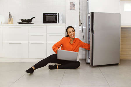 African American woman sitting sideways looking for food to eat in the refrigerator and using her hand to grab a snack while working freelance laptop in the kitchen : Women enjoy comfortable home workの写真素材