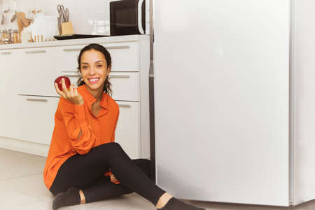 Portrait beautiful tanned woman smiling happily sitting on the floor eating apples comfortably in front of the kitchen refrigerator at home.の写真素材