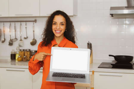 Portrait beautiful, curly-haired Latin American woman holding a laptop PC on a white background in an old rose-colored shirt, smiling happily in the modern kitchen, pointing blankly to Enter text.の写真素材