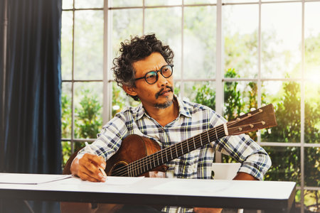 Professional Asian male composer with an acoustic guitar sat stunned, looking out the window, thinking something and wanting to concentrate, writing a song on a white sheet of paper on the table.の写真素材