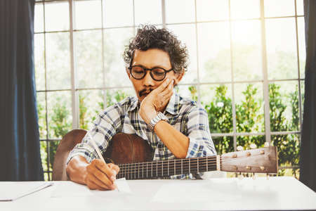 Professional Asian male composer with a wooden guitar sits obsessed with his thoughts and wants to concentrate on writing songs on paper that have no emotions.の写真素材