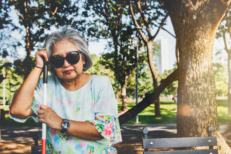 Asian woman sits in a wooden chair with a walking stick, goes blind and wears dark glasses as she sits alone in the park in the morning to calm her mind and not give up on life.の写真素材