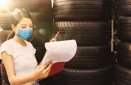 Young Asian female workers wearing masks to prevent epidemics. COVID-19 makes a list of used spare tires in the warehouse : Concept of Asian women working in establishments very seriouslyの写真素材