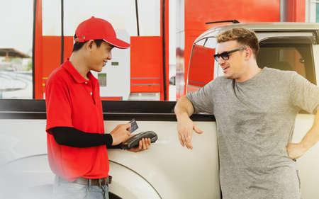 Male employee in a red uniform provides car refueling services to foreign customers and charges with...