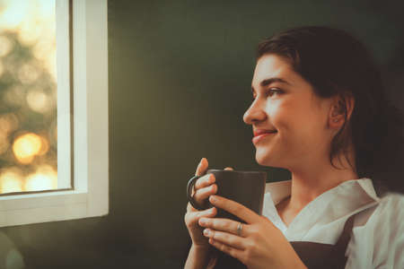 Portrait young woman barista sitting alone drinking coffee and looking out the window of a cafe with her spare time is a dreamer.の写真素材