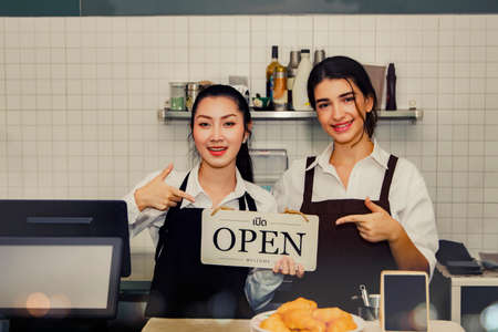 Welcome to a delicious coffee shop : Two young Asian and beautiful caucasian woman working at a coffee shop in a small coffee shop holding a smiling sign welcoming customers with a friendly smile.の写真素材